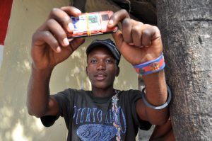 A young man wearing a Puerto Rico t-shirt is outside posing by a tree and holding his cell phone in the air as if to take a selfie.