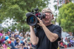 older professional photographer with cap turned backward and stubbly whiskers squints into viewfinder of large video camera in front of bokeh crowd
